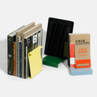 Boeken, notitieblokken en een tablet staan rechtop met de Hightide Boekenstandaard & Display Lichtblauw; op één boek zitten gele plakbriefjes. De geometrische Hightide standaard voegt stijl toe aan de effen witte achtergrond.