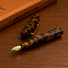 De Hightide Marmeren Fountain Vulpen Bruin van Hightide, met een gemarmerde bruine afwerking en een gouden penpunt, ligt zonder dop op een houten ondergrond naast een dicht boek met de titel ARCHITECTURE.