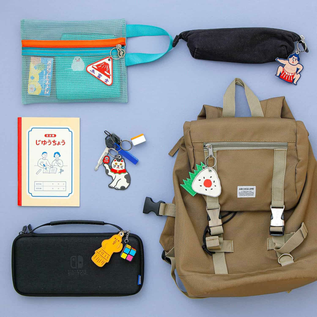 Een platte lay van een bruine rugzak, blauw etui, zwart etui, notitieblok, accessoire tas, Nintendo Switch etui en kleurrijke karakter sleutelhangers zoals de Hightide Stitch Work Sleutelhanger Fuji van Hightide op een blauwe achtergrond.