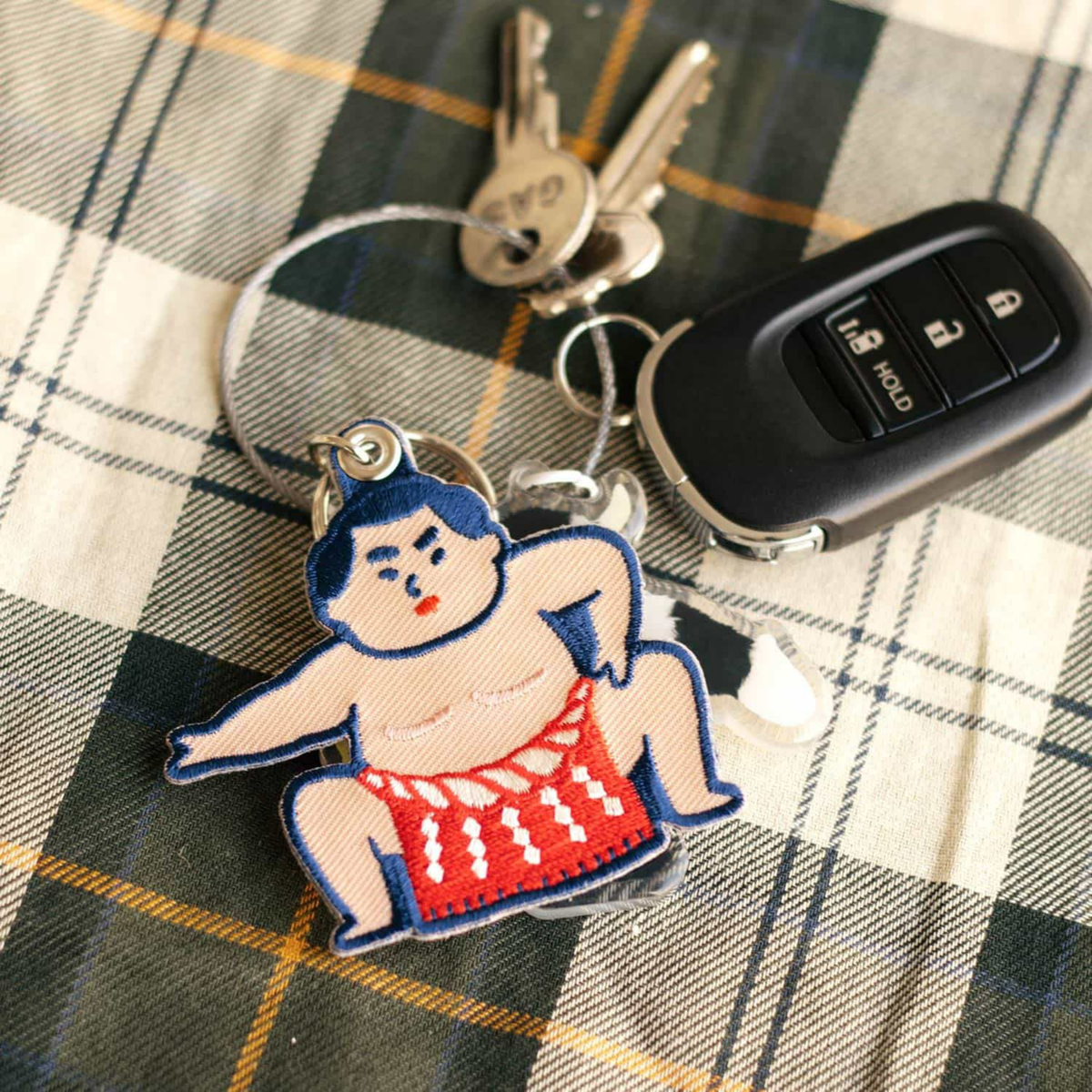 Een set sleutels, met een autosleutel en de Hightide Stitch Work Sleutelhanger Sumo Worstelaar van Hightide, met een geborduurde sumoworstelaar in een rode mawashi, zit op geruite stof.