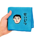 Een hand houdt de Hightide New Retro Handdoek Blauw 25/25 van Hightide vast, met een geborduurd mannengezicht en de Japanse tekst せいじつ (seijitsu), wat oprechtheid of eerlijkheid betekent.
