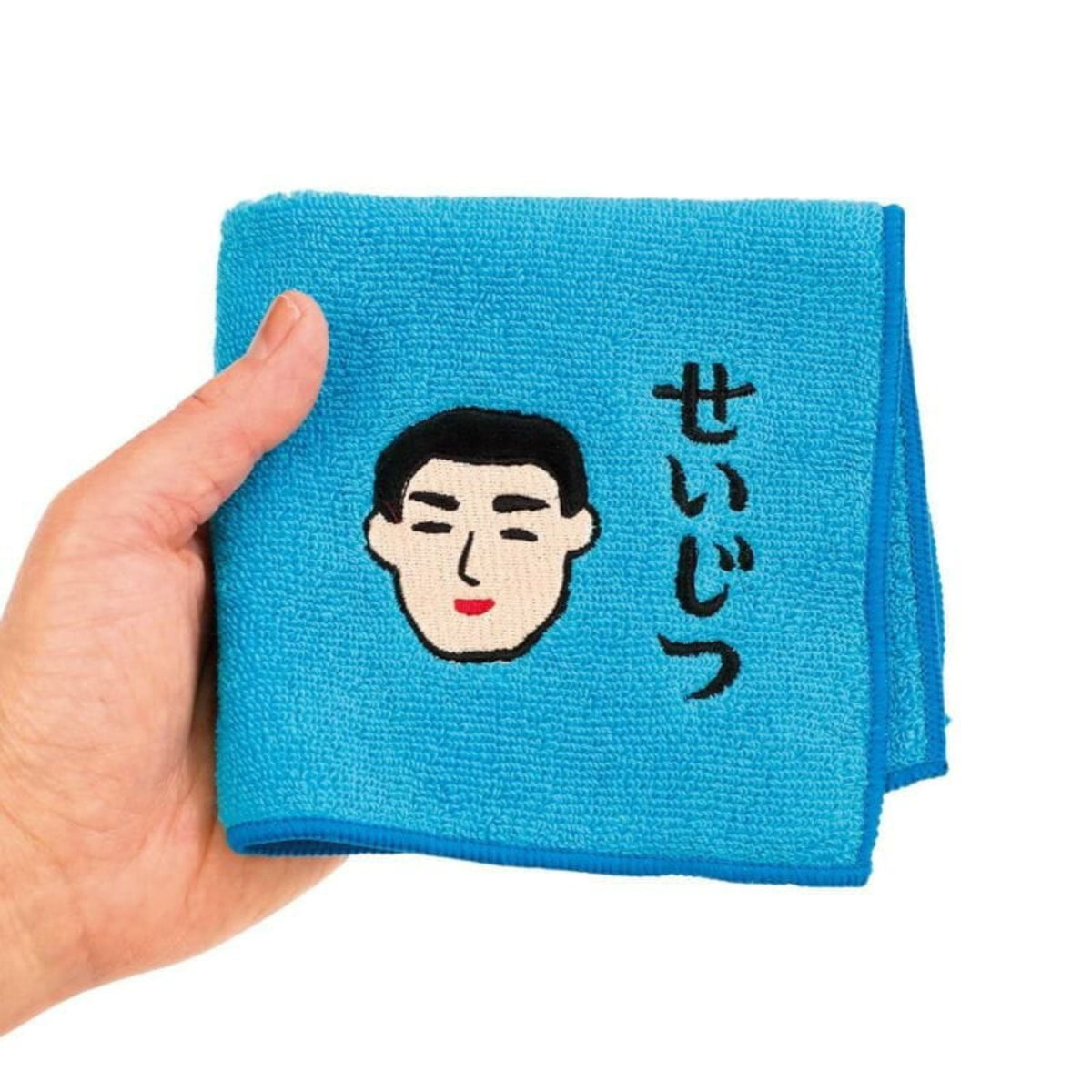 Een hand houdt de Hightide New Retro Handdoek Blauw 25/25 van Hightide vast, met een geborduurd mannengezicht en de Japanse tekst せいじつ (seijitsu), wat oprechtheid of eerlijkheid betekent.