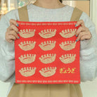 Een persoon in een grijs shirt en beige schort houdt de Hightide Handdoek Dumpling 25/25 vast, een rode handdoek van Hightide met geborduurde dumplings, decoratieve randen en "gyoza" in het Japanse hiragana in de rechterbenedenhoek.