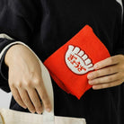 Een persoon plaatst de Hightide New Retro Handdoek Rood 25/25, een rode doek van Hightide met een witte handvormige patch, in een beige tas terwijl hij een donker shirt met lange mouwen en lichtgestreepte manchetten draagt.