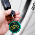 Een hand houdt een sleutel vast met de Hightide Stitch Work Sleutelhanger Fiets van Hightide, met een ronde groen geborduurde sleutelhanger met een gele fiets tegen een vaag fietswiel - ideaal voor fans van retrostijl.