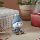 Het Hoptimist Ski Bumble S Sky beeldje, met een blauwe muts en sjaal, staat op ski's tussen ingepakte cadeautjes - een origineel cadeau en perfecte winterse decoratie met rood-wit garen en een potplant. Merk: Hoptimist.