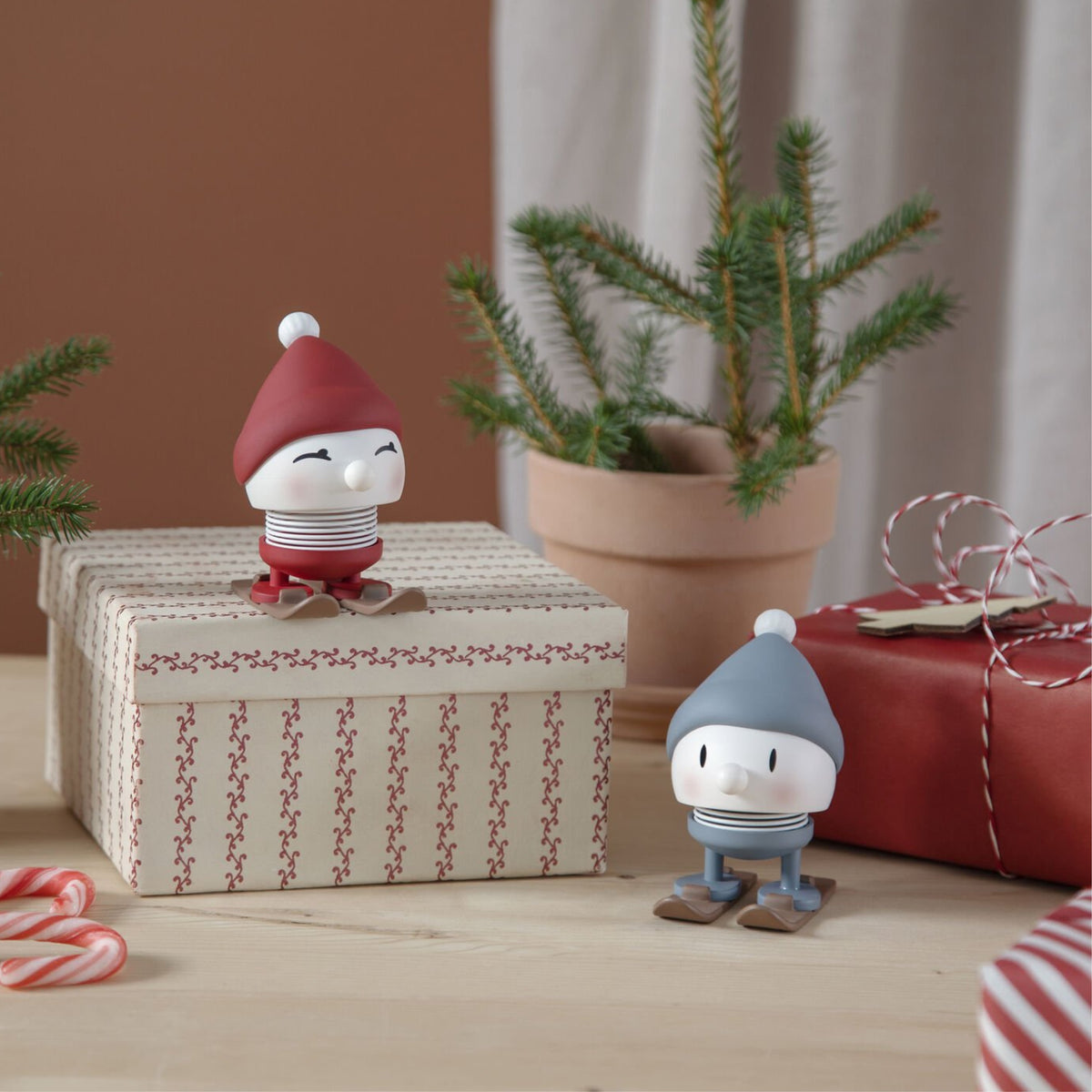 Twee beeldjes van Hoptimist Ski Bumble S Sky, met wintermutsen en sjaals, zitten op een geschenkdoos en tafel. Dit gezellige kersttafereel bevat een dennenboom in pot, zuurstokken, feestelijk koord en een origineel cadeau voor geliefden.