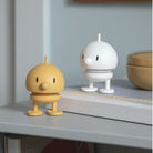 Twee kleine Hoptimist Soft Bumble S Mimosa beeldjes in geel en wit staan op gestapelde boeken en een plank naast een bijpassende schaal in een minimale, moderne kamer geïnspireerd door Deens design.