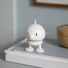 De Hoptimist Soft Bumble S Wit, een klein, wit beeldje van Hoptimist met een groot hoofd en ronde neus, staat op een dienblad met rasterpatroon naast een beige bakje tegen een lichtblauwe achtergrond, geïnspireerd op Deens design.