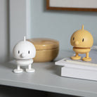 Een wit Hoptimist Soft Bumble S beeldje van Hoptimist staat op een plank naast een gesloten notitieboekje en een beige container en voegt speels Deens design toe aan de scène tegen een lichte muur met ingelijste foto.
