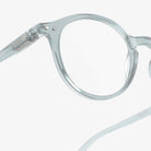 Close-up van transparant, rond Izipizi Leesbril #D Frozen Blue montuur tegen een witte achtergrond, met de linker lens en een deel van de veerarm van Izipizi, ontworpen voor comfortabele correctie van ouderdomsverziendheid.