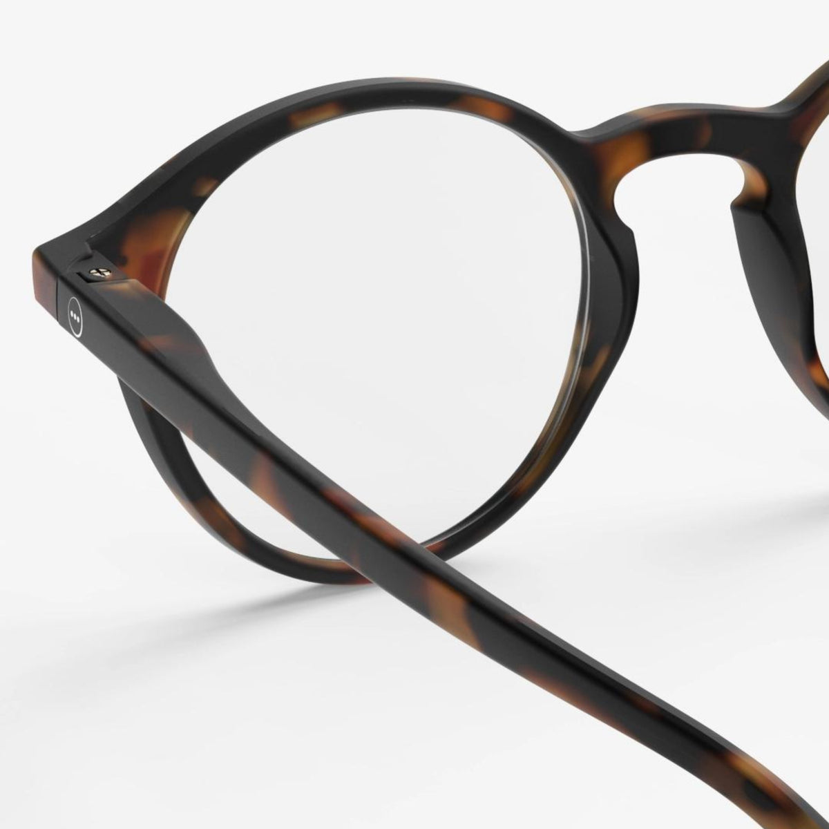 Een close-up van de Izipizi Leesbril #D Tortoise van Izipizi, met een ronde bril met een donker tortoiseshell montuur, met de focus op de linker lens en arm tegen een effen witte achtergrond.