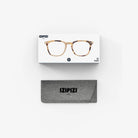 De Izipizi Leesbril #E Light Tortoise van Izipizi, met een stijlvol schildpadmontuur, wordt weergegeven boven een grijze merketui op een witte achtergrond-ideaal als een modieuze leesbril heren dames voor dagelijks lezen.