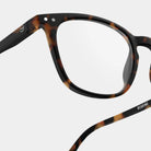 Close-up van de Izipizi Leesbril #E Tortoise leesbril van Izipizi met heldere glazen op een effen achtergrond. "READING" staat aan de binnenkant van het pootje en een witte smiley siert het scharnier. Stijlvolle unisex leesbril voor dagelijks gebruik.