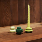 Drie ronde &Klevering Bravo Kaarsen Groen kandelaars van &Klevering, in lichtgeel en groen, zitten op hout; één houdt een gedraaide lichtgele kaars vast, de andere zijn leeg. Ideaal voor kleurrijke kaarsen of om cadeau te geven in een luxe geschenkdoos.