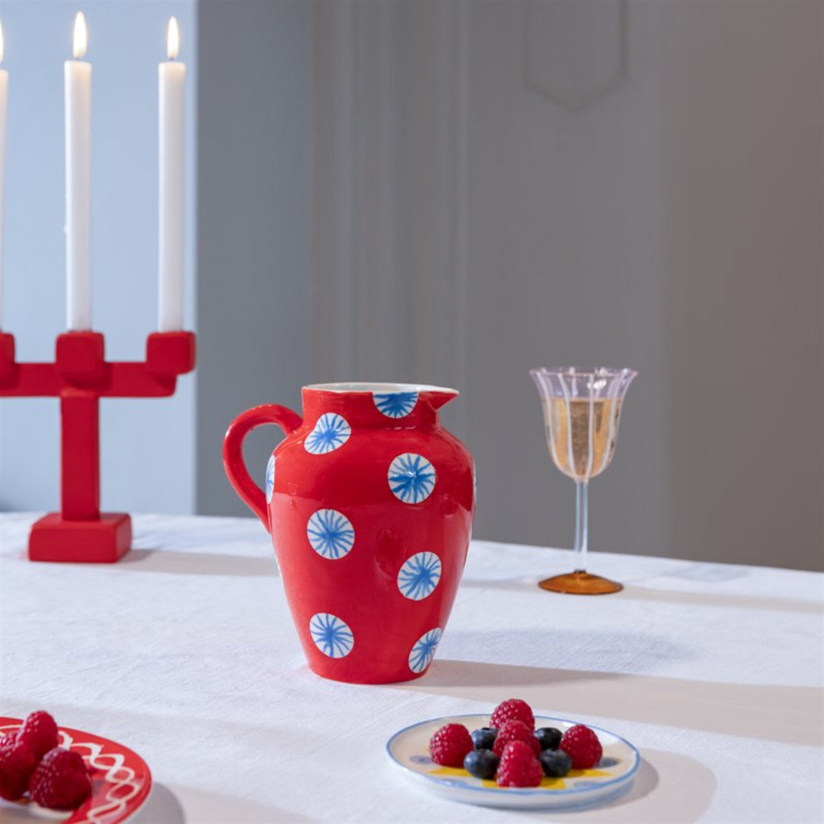 Een &Klevering Festivo Kan Rood 1,5L met blauwe en witte cirkels rust op een wit tafelkleed naast een bord bessen, een champagneglas en een rode kandelaar met drie brandende kaarsen, die samen een charmante decoratieve display vormen.