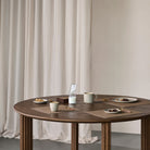 Een ronde houten tafel gedekt met Lind DNA Serene Placemat Rechthoekig Molgrijs 35/45, kleine snackbordjes, kopjes, een mok, gouden lepels, een melkkaraf en een doos staat opgesteld voor beige gordijnen.