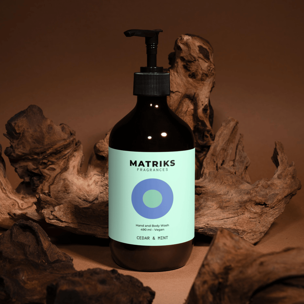 Een 490 ml fles Matriks Fragrances Hand & Body Wash Cedar & Mint met pompje staat tussen drijfhout op een bruine achtergrond.