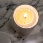 Een brandende Matriks Fragrances Geurkaars Keramiek Woodland Pine 270 g staat in zijn witte pot op marmer. De stabiele vlam gloeit, handgemaakt in Antwerpen, met het deksel van de kaars dichtbij.
