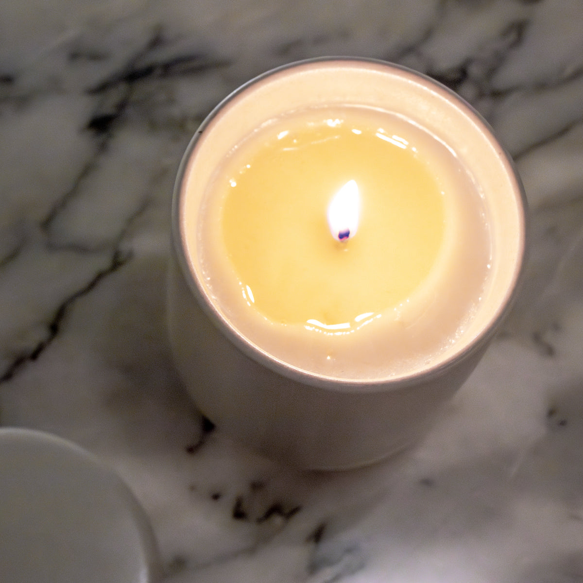 Een brandende Matriks Fragrances Geurkaars Keramiek Woodland Pine 270 g staat in zijn witte pot op marmer. De stabiele vlam gloeit, handgemaakt in Antwerpen, met het deksel van de kaars dichtbij.