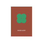 De Matriks JL Postkaart Good Luck Dubbel van Matriks heeft een bruine achtergrond, een groen klavertjevier-illustratie in het midden en "GOOD LUCK!" eronder geschreven in witte hoofdletters.