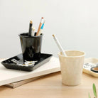 Een gemarmerde Melamine Beker Zwart en een beige beker, beide gevuld met pennen en potloden, rusten op een houten bureau naast een organizer tray met paperclips en scharen, tegen een minimalistische neutrale achtergrond.