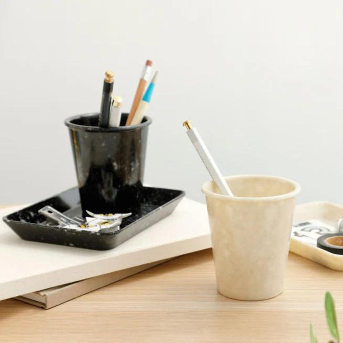 Een gemarmerde Melamine Beker Zwart en een beige beker, beide gevuld met pennen en potloden, rusten op een houten bureau naast een organizer tray met paperclips en scharen, tegen een minimalistische neutrale achtergrond.