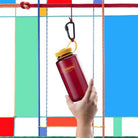 Een hand houdt een rode Nalgene Drinkfles Brede Opening Laker 1L met gele dop, bevestigd aan een karabijnhaak. Op de achtergrond zijn kleurrijke geometrische vormen in blauw, rood, groen en wit zichtbaar.
