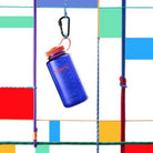 De Nalgene Drinkfles Brede Opening Periwinkle 1L van Nalgene, met een oranje deksel, hangt aan een karabijnhaak tussen kleurrijke touwen tegen een geometrische veelkleurige achtergrond.