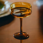 Een close-up van de Paveau Swirl Champagne Coupe Dune Geel 26 cl van Paveau, gemaakt van borosilicaatglas, staat leeg op een houten tafel met zacht licht dat weerkaatst op het amberkleurige oppervlak.