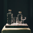 Twee Paveau Swirl Waterfles Wave Helder 0,75 l flessen met witte doppen, drie lege glazen en een coupeglas staan bovenop een groot boek met de titel Gert Voorjans op een lichtgroen oppervlak tegen een donkere achtergrond.