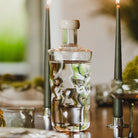 De Paveau Swirl Waterfles Wave Helder 0,75 l van Paveau met witte dop en textuur staat op een houten eettafel naast twee brandende kaarsen, met een vage groene achtergrond.