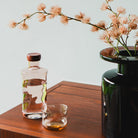 De Paveau Swirl Waterfles Glow Beige 1,25 l van Paveau staat op een houten ondergrond naast een glas water en een vaas met lichtroze bloemen - een ideaal duurzaam cadeau tegen een lichte achtergrond.