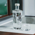 Een glazen karaf met een witte stop staat naast een Paveau Swirl Waterglas Wave Helder 24 cl van Paveau, beide gemaakt van duurzaam glas en geplaatst op een aanrecht in de buurt van een spiegel.