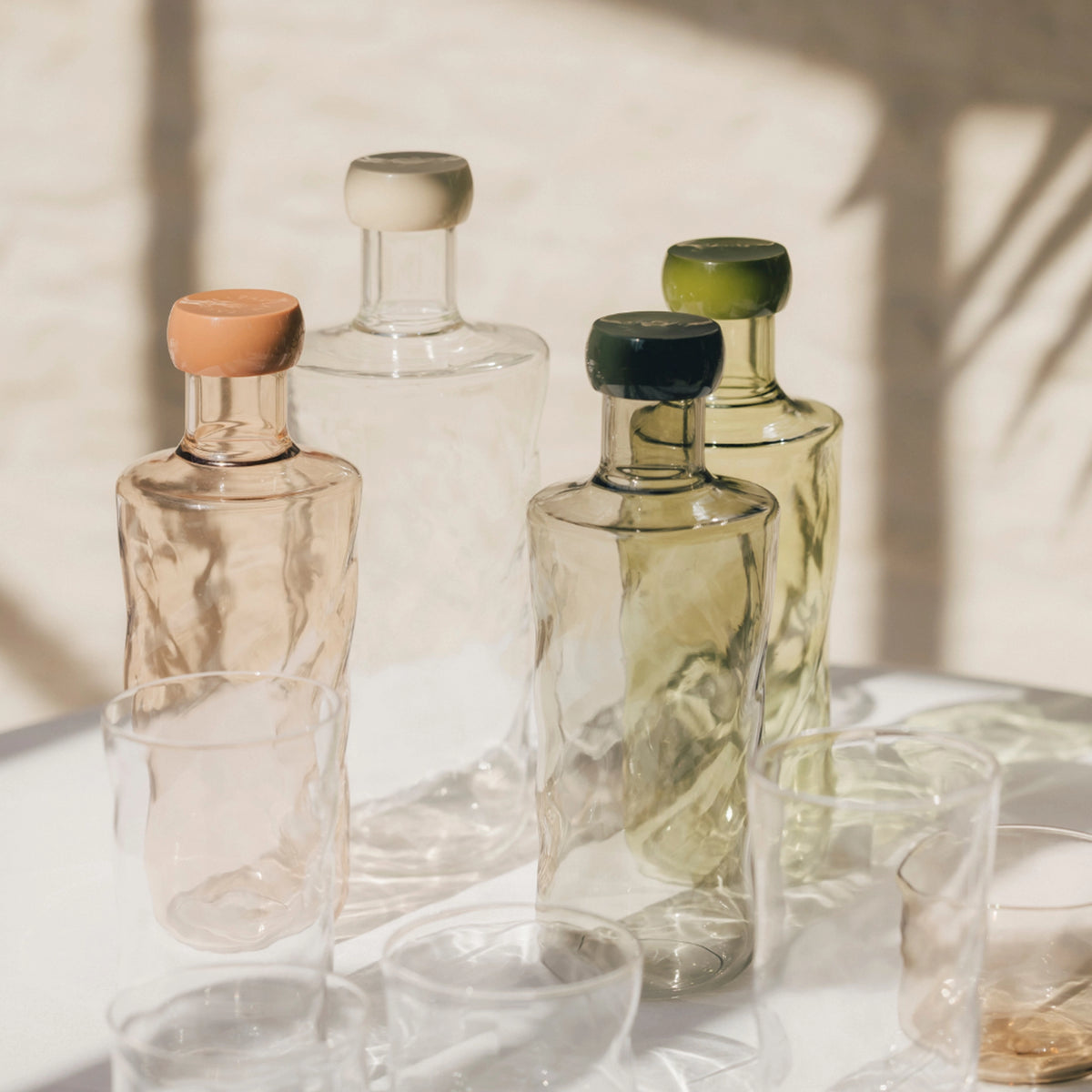 De Paveau Swirl Waterfles Moss Groen 0,75 l van Paveau, met golvend design en pastelgroene dop, staat samen met bijpassende borosilicaatglas tumblers zacht belicht op een zonovergoten witte ondergrond.