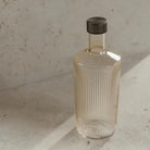 De Paveau Waterfles Bells Beige 1,25 l van Paveau heeft een helder, geribbeld ontwerp met een donkere kap, rustend op een licht stenen oppervlak. Zonlicht versterkt de minimalistische en elegante stijl.