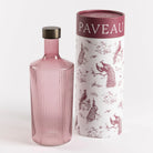Een geribbelde, roze glazen Paveau Waterfles Broome 1,25 l met een donkere dop staat naast een witte cilindrische doos versierd met bordeauxrode pauwen en bomen, met "PAVEAU" bovenaan.