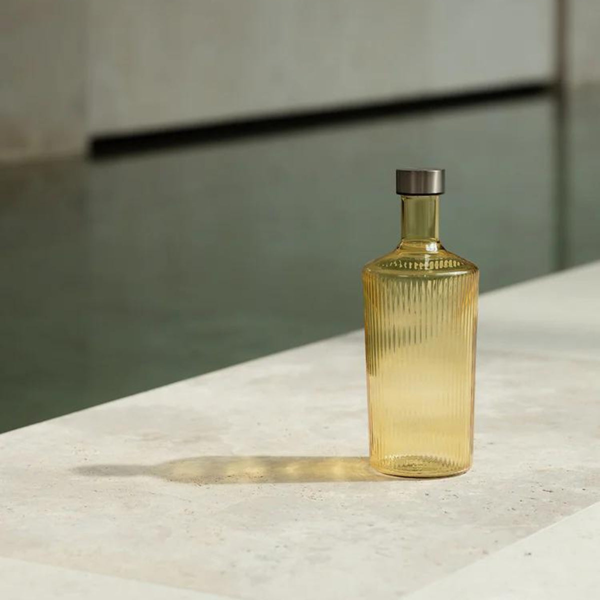De Paveau Waterfles Cable Yellow 1,25 l van Paveau, met een geribbeld amberkleurig design en metalen kap, rust op een licht stenen oppervlak met een lange schaduw; water en neutrale muren vervagen op de achtergrond.