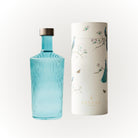 Een Paveau Waterfles Lucky Turquoise 1,25 l met een metalen dop staat naast een hoge witte cilindrische doos versierd met bloemmotieven, vlinders en pauwen. Op de bodem van de doos staat het merk Paveau.