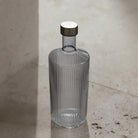 De Paveau Waterfles Twilight Blue 1,25 l van Paveau, met een helder geribbeld ontwerp en metalen kap, staat rechtop op een beige stenen oppervlak dat baadt in zacht natuurlijk licht.