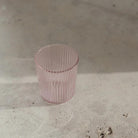 De Paveau Waterglas Pink van Paveau, een lichtroze geribbelde glazen beker, staat leeg op een licht gestructureerd oppervlak in zacht natuurlijk licht, en werpt een subtiele schaduw.