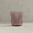 De Paveau Waterglas Broome van Paveau, met een roze tint en verticaal geribbeld ontwerp, staat op een zacht neutraal oppervlak - ideaal voor iedereen die op zoek is naar een stijlvolle, duurzame drinkbril.
