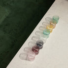 Acht geribbelde, doorschijnende Paveau Waterglas Broome by Paveau in verschillende kleuren staan diagonaal opgesteld op een licht oppervlak met zachte schaduwen, naast een donkergroene tegelvloer.