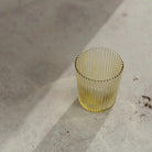 De Paveau Waterglas Cable Yellow, een transparant geribbeld geel glas van Paveau, ligt op een licht oppervlak en werpt een zachte schaduw in het zonlicht. Ontdek de duurzame Paveau waterglas voor elke gelegenheid.