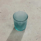 De Paveau Waterglas Lucky Turquoise van Paveau rust op een lichtbeige gestructureerd oppervlak. Het transparante, geribbelde groenblauwe glas werpt een zachte schaduw naar links.