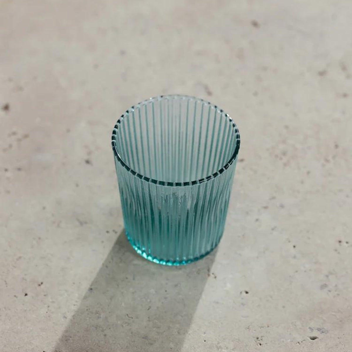 De Paveau Waterglas Lucky Turquoise van Paveau rust op een lichtbeige gestructureerd oppervlak. Het transparante, geribbelde groenblauwe glas werpt een zachte schaduw naar links.