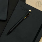 Een Penco 4 Kleuren Balpen Zwart met vintage industriële look en gouden clip ligt schuin op een gesloten zwart notitieboek, terwijl een deel van een lichtbruine envelop en een zwart object op de achtergrond zichtbaar zijn.
