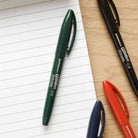 Een Penco Glider Pen Blauw ligt diagonaal op een gelinieerd notitieblok en zorgt voor een zijdezachte schrijfervaring, met zwarte, groene en rode Penco pennen in de buurt op een houten oppervlak.