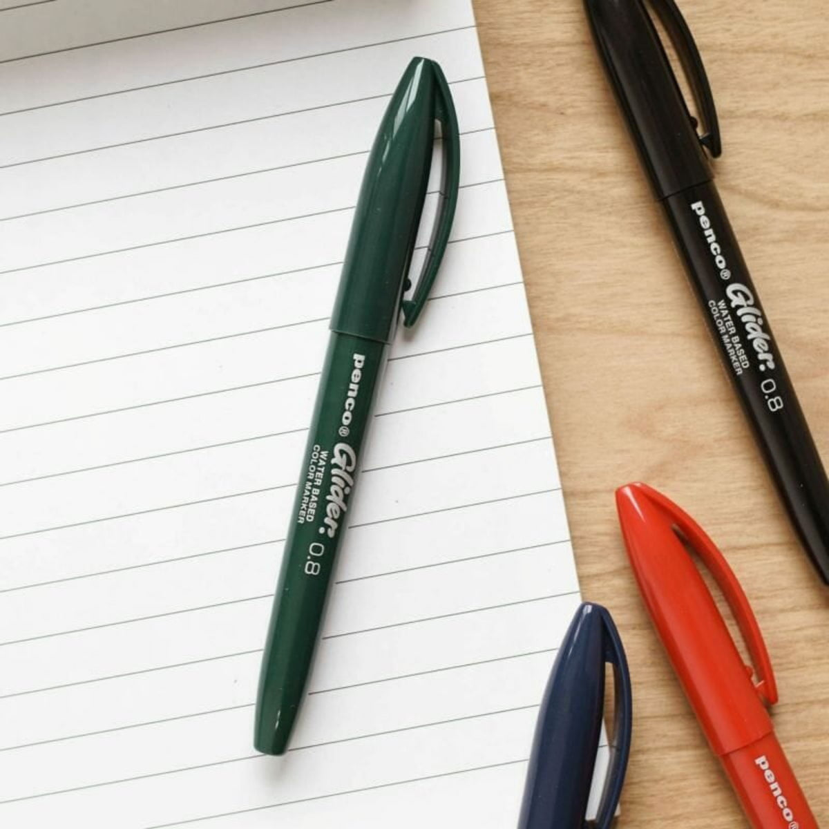 Een Penco Glider Pen Blauw ligt diagonaal op een gelinieerd notitieblok en zorgt voor een zijdezachte schrijfervaring, met zwarte, groene en rode Penco pennen in de buurt op een houten oppervlak.