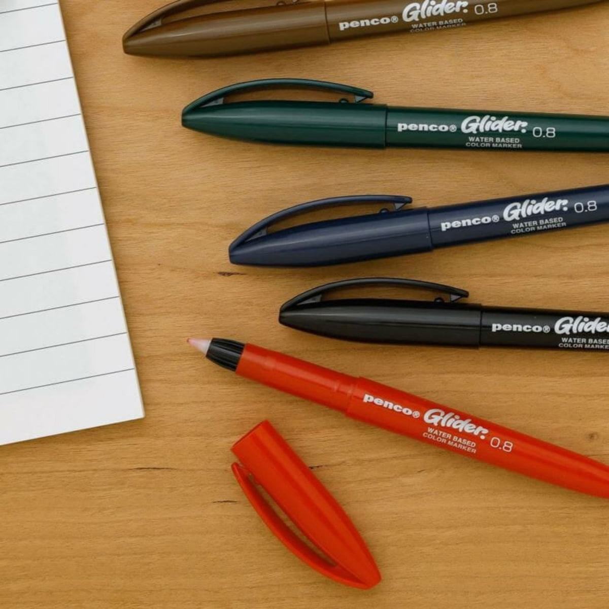 Vier Penco Glider Pennen Blauw 0,8 (bruin, groenblauw, marineblauw en rood) liggen op een houten oppervlak naast een gelinieerd notitieblok. De rode pen zonder dop toont de punt voor een zijdezachte schrijfervaring.